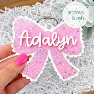 May include: A pink glittery bow-shaped keychain with a white scalloped border and the name Adalyn in raised white script lettering. The item features a silver metal keyring attachment at the top for securing to bags or other personal items.