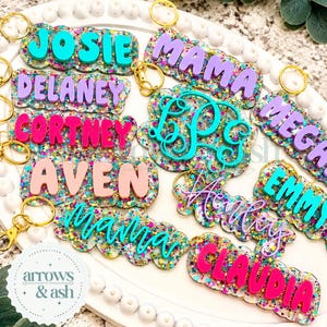 May include: A collection of custom acrylic keychains featuring scalloped edges and multi-colored glitter backgrounds. Each keychain displays a name or monogram in raised, solid-colored lettering, including Josie, Mama, Delaney, Cortney, Aven, Henley, Emmy, and Claudia.