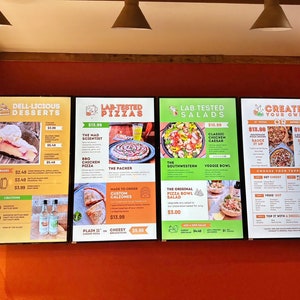 May include: Three digital menu boards with a red brick background. The first board lists desserts, the second lists lab-tested pizzas, and the third lists lab-tested salads. The fourth board has a create your own pizza menu with toppings and sauces.