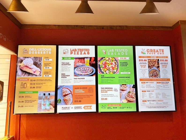 May include: Three digital menu boards with a red brick background. The first board lists desserts, the second lists lab-tested pizzas, and the third lists lab-tested salads. The fourth board has a create your own pizza menu with toppings and sauces.