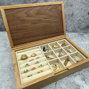 May include: Wooden jewellery box with a light interior and several compartments. The box is filled with rings of various designs, including gold and silver rings, some with gemstones. The lid is open, revealing a dark wood grain.