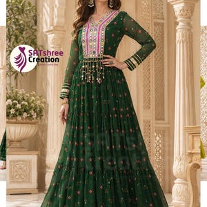 May include: A floor-length dark green dress with long sleeves, featuring a V-neckline adorned with pink and silver embroidery, reflective mirror accents, and dangling metallic charms at the waist. The fabric has a small repeating geometric pattern.