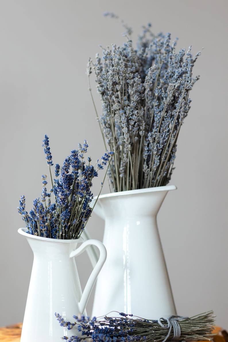 May include: Two white ceramic pitchers with dried lavender flowers. The pitchers are on a wooden surface.