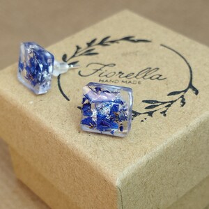 Könnte beinhalten: Ein Paar quadratische Harzohrringe mit gepressten blauen Blumen und silbernen Flocken im Inneren. Die Ohrringe befinden sich in einer kleinen braunen Schachtel mit dem Text "Fiorella Hand Made" darauf.