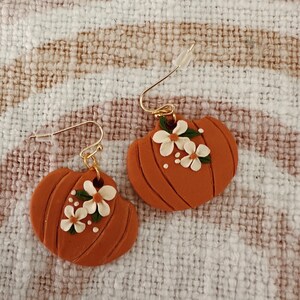 May include: A pair of orange pumpkin earrings with gold-colored hooks. Each earring features a pumpkin shape with carved lines and small white flowers with green leaves. The earrings are set against a textured, off-white and brown background.