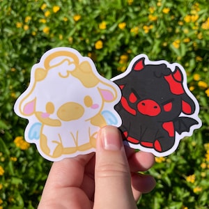 May include: Two stickers featuring cartoon cows. One is a white cow with yellow accents, angel wings, and a halo. The other is a black cow with red accents, horns, and bat wings. The stickers are held against a background of yellow flowers.