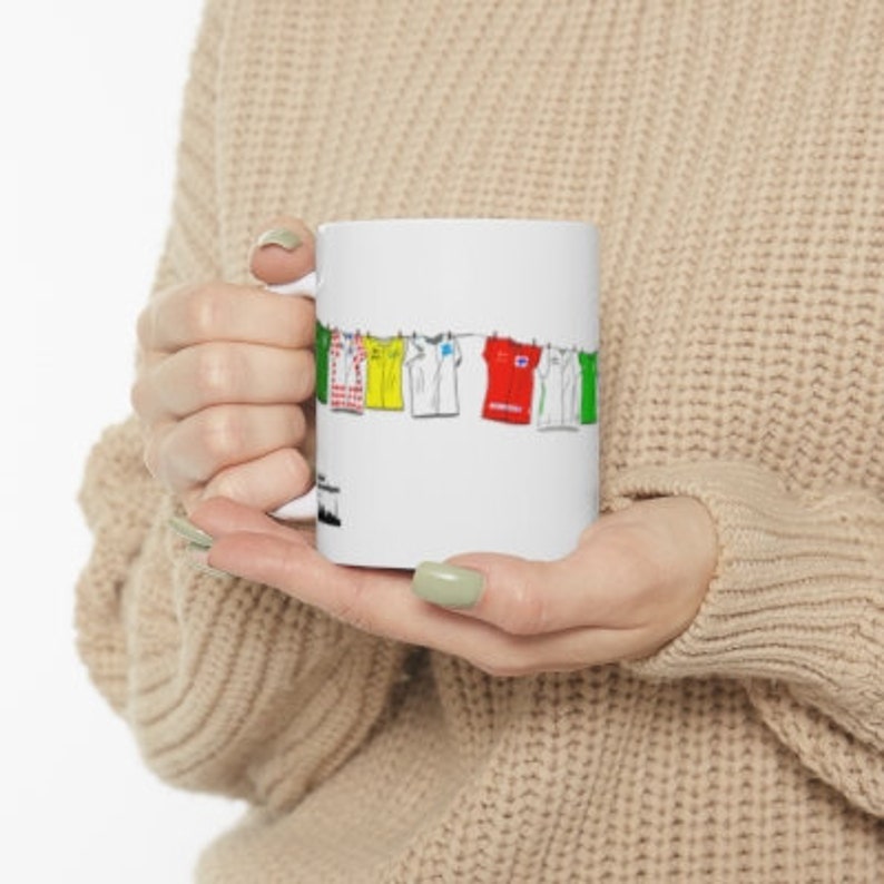 May include: White ceramic mug with a colorful illustration of clothes hanging on a clothesline. The clothes are yellow, red, green, blue, and white.