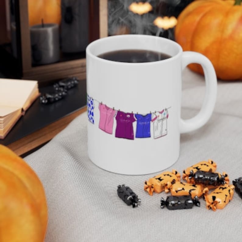 May include: White ceramic coffee mug with a design of five colorful shirts hanging on a clothesline. The shirts are blue, pink, purple, blue, and white.