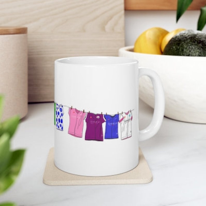 May include: White ceramic mug with a line of colorful shirts hanging on a clothesline printed on the side. The shirts are green, pink, purple, blue, and white.