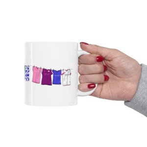 May include: White ceramic mug with a colorful illustration of clothes hanging on a clothesline. The clothes are a green and white patterned shirt, a pink shirt, a purple shirt, a blue shirt, and a white shirt.