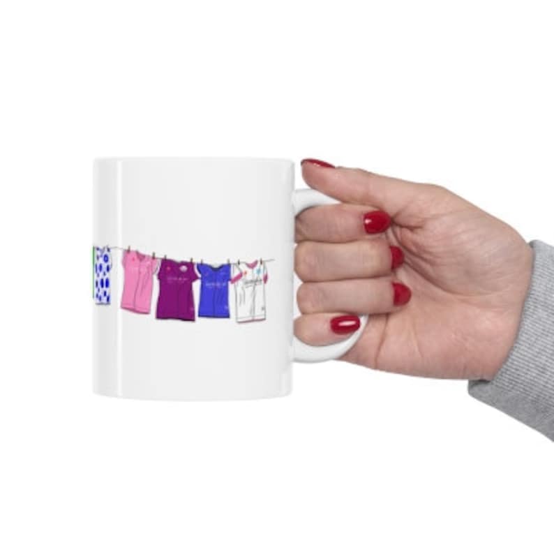 May include: White ceramic mug with a colorful illustration of clothes hanging on a clothesline. The clothes are a green and white patterned shirt, a pink shirt, a purple shirt, a blue shirt, and a white shirt.