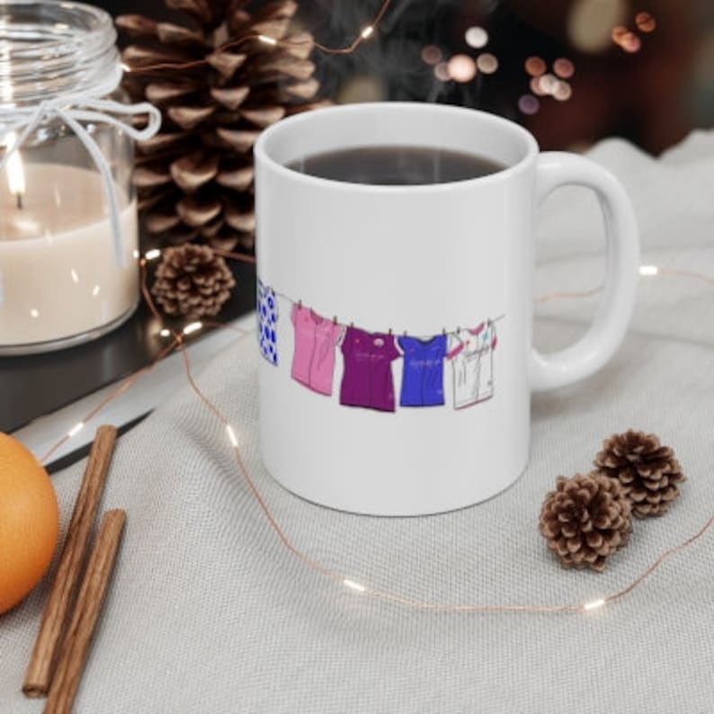 May include: White ceramic mug with a graphic of five colorful shirts hanging on a clothesline. The shirts are blue, pink, purple, blue, and white.