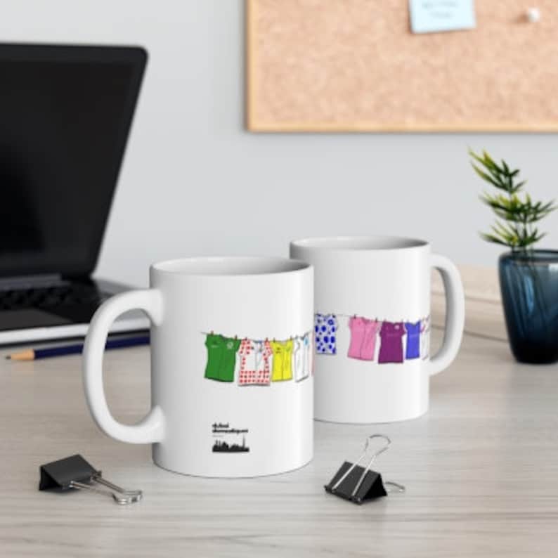 May include: Two white ceramic mugs with a design of colorful shirts hanging on a clothesline. The text "Official Tour de France" is printed on the mug on the left.