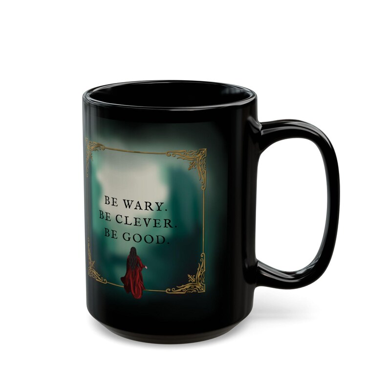 May include: Black ceramic mug with a curved handle. The mug features the text "BE WARY. BE CLEVER. BE GOOD." in white, framed by a gold decorative border. A figure in a red cloak is also depicted.