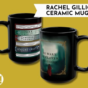 May include: Two black ceramic mugs. One mug features a stack of books with titles like "The Dark Window" and "Twisted Crowns." The other mug has the text "Be wary. Be clever. Be good." with a figure in a red cloak. The mugs are labeled "RACHEL GILLIG CERAMIC MUG!"