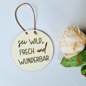 Può includere: Un cartello rotondo in legno chiaro con la scritta "sei WILD, FRECH und WUNDERBAR" in corsivo nero. Il cartello ha un cordino in pelle marrone per appenderlo. Sullo sfondo, una rosa color crema con foglie verdi.