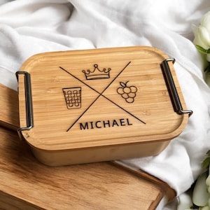 Könnte beinhalten: Eine rechteckige Bambus-Lunchbox mit Deckel, verziert mit einer Krone, einem Glas und Trauben, darunter der Name "MICHAEL". Die Box hat Metallverschlüsse und steht auf einer Holzoberfläche.
