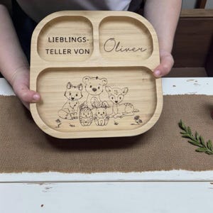 Op de afbeelding: Een houten verdeelde plaat met gegraveerde illustraties van bosdieren. De plaat heeft drie secties en bevat de naam "Oliver" en de tekst "LIEBLINGS-TELLER VON". De plaat is gemaakt van lichtgekleurd hout.