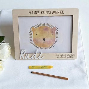 Op de afbeelding: Houten fotolijst met de tekst "MEINE KUNSTWERKE" bovenaan. Binnenin een aquarel illustratie van een leeuw. De naam "Rudi" staat eronder. Een gele krijt en een bruine stift liggen eronder.