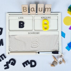 Può includere: Un giocattolo di apprendimento in legno con un vassoio di sabbia per scrivere la lettera 'B'. Il giocattolo ha due sezioni con le parole 'Lies' e 'Kreierre' e una sezione con la parola 'Schreibe'. Il giocattolo include anche tessere di lettere in legno che compongono 'Baum' e tessere di lettere in schiuma gialla che compongono 'B'.