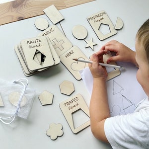 Può includere: Un bambino traccia una forma con una matita su un foglio di carta bianco.  Forme di legno con nomi tedeschi e ritagli sono sparse su un tavolo bianco.  Le forme includono un cuore, una stella, una croce, un rombo, un trapezio e un pentagono.