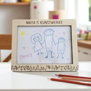 Op de afbeelding: Houten fotolijst met de tekst "MAYA'S KUNSTWERKE". Binnenin is een familietekening met het woord "FAMILY" eronder. De onderkant van de lijst heeft een bloemenpatroon. Er liggen verschillende rode potloden op de tafel.