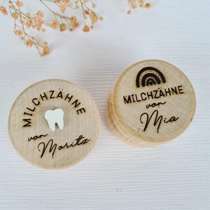 Op de afbeelding: Twee houten dozen met deksels om babytanden in te bewaren. Eén doos heeft een tandsymbool en de tekst "MILCHZÄHNE von Morita". De andere doos heeft een regenboogicoon en de tekst "MILCHZÄHNE von Mia".