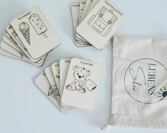 May include: A set of wooden flash cards with various images and German words. The cards are stacked in piles and include images of a teddy bear, ice cream, and a backpack. The cards are on a white surface. The text on the cards includes "Eis essen", "Kuscheln", and "Tasche packen".