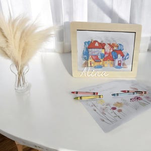 Könnte beinhalten: Ein aquarelliertes Bild von bunten Häusern in einem Holzrahmen mit roten, gelben und blauen Dächern. Der Name "Alina" steht darunter. Buntstifte und eine Blumenskizze liegen auf dem Tisch.