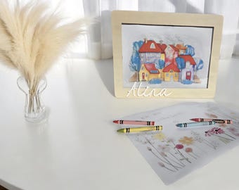 Può includere: Un dipinto ad acquerello incorniciato di case colorate con tetti rossi, gialli e blu. Il nome "Alina" è scritto sotto. Ci sono pastelli e un disegno floreale sul tavolo.
