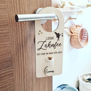 Könnte beinhalten: Ein hölzerner Türhänger mit dem Text "Liebe Zahnfee" und "Ich hab da was fur dich". Eine kleine Glasflasche mit einem Korkverschluss ist am Hänger befestigt. Die Flasche ist mit "Emmy" beschriftet.
