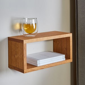 Könnte beinhalten: Ein schwebendes Holzregal mit einem Glas Tee und einem Stapel Bücher darauf. Das Regal ist an einer weißen Wand neben einem grauen, gepolsterten Kopfteil befestigt.