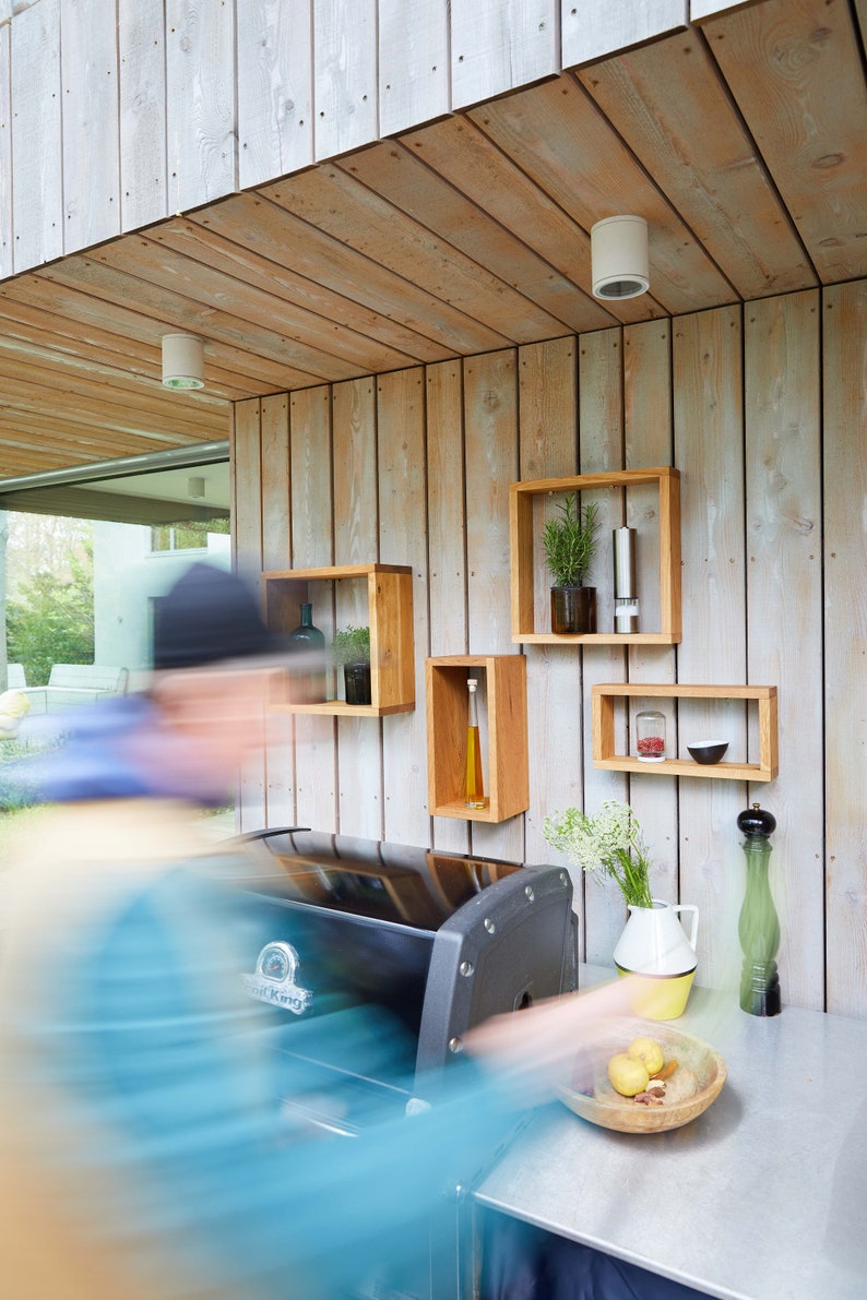 K&ouml;nnte beinhalten: Ein schwarzer Outdoor-Grill mit einer Arbeitsplatte aus Edelstahl und Holzregalen. Die Regale sind mit Kr&auml;utern, Gew&uuml;rzen und &Ouml;l gef&uuml;llt. Auf der Arbeitsplatte steht eine Sch&uuml;ssel mit Obst.