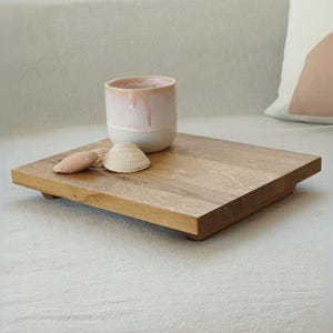 Könnte beinhalten: Ein helles Holztablett mit abgerundeten Kanten und vier kleinen Füßen. Das Tablett befindet sich auf einer weißen Oberfläche und hat eine rosa-weiße Keramiktasse und zwei Muscheln darauf.