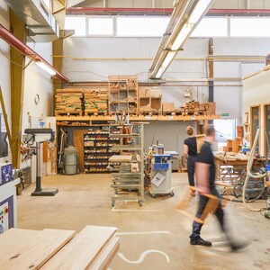 K&ouml;nnte beinhalten: Eine gro&szlig;e Werkstatt mit Holz und Werkzeugen. Eine Person steht in der Mitte des Raumes und tr&auml;gt ein blaues Hemd und Jeans. An den W&auml;nden befinden sich Regale mit Holz und Werkzeugen, und im Vordergrund steht eine gro&szlig;e Maschine.