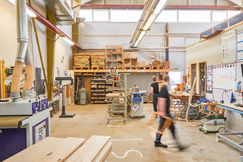K&ouml;nnte beinhalten: Eine gro&szlig;e Werkstatt mit Holz und Werkzeugen. Eine Person steht in der Mitte des Raumes und tr&auml;gt ein blaues Hemd und Jeans. An den W&auml;nden befinden sich Regale mit Holz und Werkzeugen, und im Vordergrund steht eine gro&szlig;e Maschine.