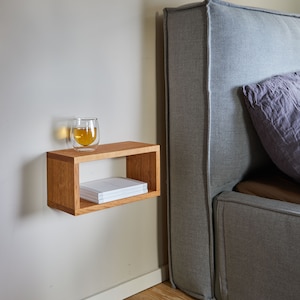 K&ouml;nnte beinhalten: Ein schwebendes Holzregal mit einem Glas Tee und einem Stapel B&uuml;cher darauf. Das Regal ist an einer hellbeigen Wand neben einem grauen, gepolsterten Kopfteil befestigt.