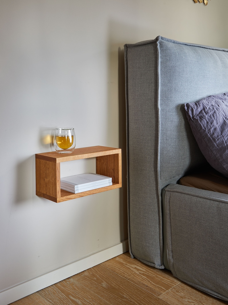 K&ouml;nnte beinhalten: Ein schwebendes Holzregal mit einem Glas Tee und einem Stapel B&uuml;cher darauf. Das Regal ist an einer hellbeigen Wand neben einem grauen, gepolsterten Kopfteil befestigt.