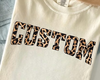 May include: Cream-colored t-shirt with the word "CUSTOM" in a leopard print design. The shirt is folded and placed on top of a pair of blue jeans and a tan hat.