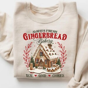 May include: Beige sweatshirt featuring a gingerbread house design with the text "ALWAYS FRESH GINGERBREAD Bakery". The design includes red accents and the words "REAL GOOD COOKIES". The sweatshirt is folded.