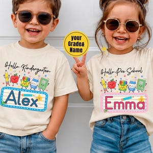 Pu&ograve; includere: Due magliette beige con grafiche colorate a tema scolastico tra cui una matita, una mela, pastelli, un cartone di latte e un quaderno. La maglietta a sinistra mostra il testo Hello Kindergarten e Alex, quella a destra Hello Pre School e Emma.