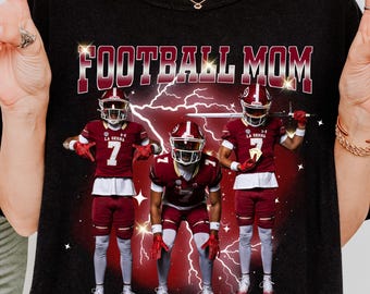 May include: Black t-shirt with the text "FOOTBALL MOM" in a bold, outlined font. The graphic features three football players in maroon and white uniforms with lightning bolts in the background. The players are wearing helmets and pads.