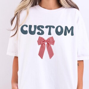 May include: White t-shirt with the word "CUSTOM" in teal, above a red and white gingham bow graphic. The shirt has short sleeves and a relaxed fit.