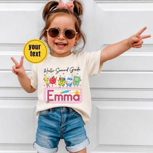 Pu&ograve; includere: Una t-shirt a maniche corte color crema con la scritta Hello Second Grade sopra disegni animati di materiale scolastico tra cui una matita, una mela, pastelli, un cartone di latte e un quaderno, con il nome Emma in un riquadro rosa.
