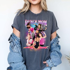 K&ouml;nnte beinhalten: Ein dunkelgraues T-Shirt mit einem grafischen Aufdruck einer Gruppe tanzender M&auml;dchen. Der Text "Dance Mom Kate" ist auf dem Shirt aufgedruckt.