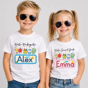 Pu&ograve; includere: Due magliette bianche a maniche corte con grafiche colorate di articoli scolastici e testo. La maglietta a sinistra riporta Hello Kindergarten e Alex, quella a destra Hello Second Grade e Emma. Entrambe presentano disegni di matita, mela, pastelli, latte e quaderno.