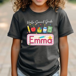 Pu&ograve; includere: Una t-shirt grigio scuro con la scritta Hello Second Grade e una grafica raffigurante una matita, una mela, pastelli, un cartone di latte e un quaderno. Sotto, un rettangolo rosa contiene il nome Emma con una piccola matita e una stella.