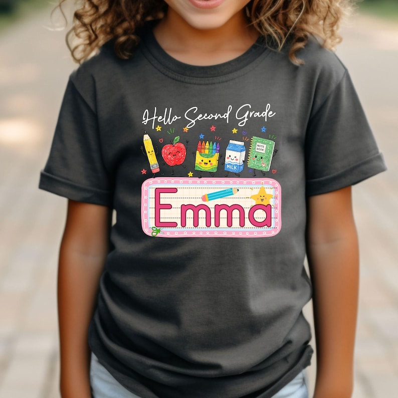 Pu&ograve; includere: Una t-shirt grigio scuro con la scritta Hello Second Grade e una grafica raffigurante una matita, una mela, pastelli, un cartone di latte e un quaderno. Sotto, un rettangolo rosa contiene il nome Emma con una piccola matita e una stella.
