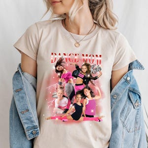 K&ouml;nnte beinhalten: Ein beiges T-Shirt mit einem Grafikdruck von f&uuml;nf tanzenden M&auml;dchen. Die Grafik enth&auml;lt den Text "Dance Mom Kate" in rosa und wei&szlig;en Buchstaben.