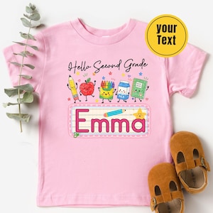 Pu&ograve; includere: Una t-shirt a maniche corte rosa chiaro con una grafica che include il testo Hello Second Grade ed Emma. Il design presenta illustrazioni a cartoni animati di una matita, una mela, pastelli, un cartone di latte, un quaderno e una stella, tutti con facce sorridenti.